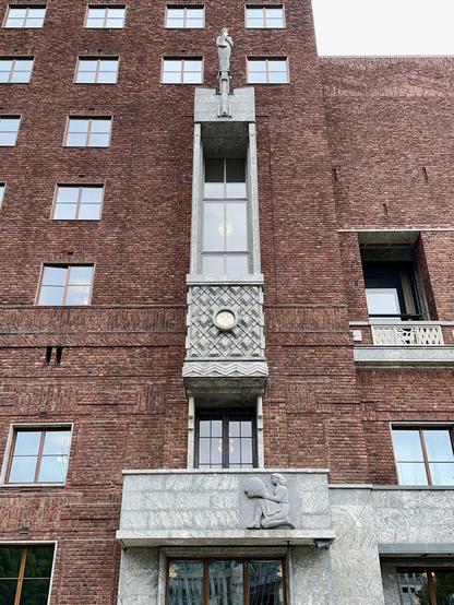 Image of a building facade featuring red brick walls, large windows, and decorative granite elements. There is a prominent sculpture of a figure standing atop a pillar, as well as a clock and a relief of a kneeling figure holding an object.