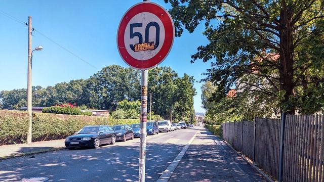Ein Tempo 50 Schild in der Bildmitte. Dahinter eine Straße. Links parken Autos. Rechts ein breiter Fußweg, der aber leicht überfahren werden kann. Dazwischen eine Straße, die etwas knapp ist für zwei Fahrspuren.