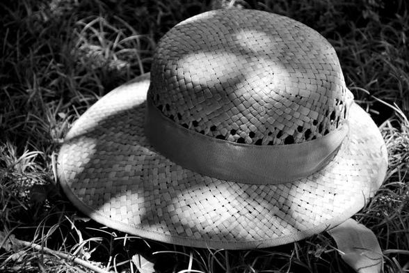 C'est une photo en noir et blanc. Sur fond d'herbe un peu sèche, un chapeau de paille à large bord, avec un ruban et un motif de tressage ménageant une aération, occupe presque la totalité de l'image.  Le soleil est filtré par un feuillage, ce qui fait un jeu d'ombre et de lumière sur la paille du chapeau.