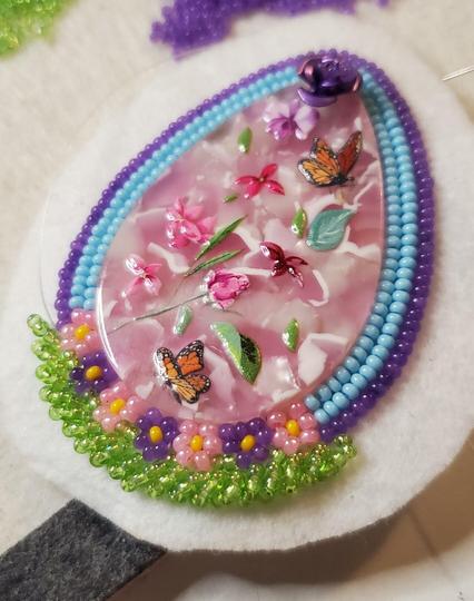 the beginnings of a beaded teardrop shaped earring made by kiamichi. the centerpiece is made of pink enamel with painted decorations of roses, flowers, and butterflies. that piece is surrounded by beaded pink and purple flowers with green underneath and bordered by blue and purple