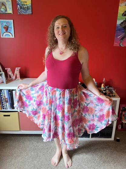 miriam, a white trans woman with long curly dirty blonde hair, in a raspberry pink tank top and pink, white, and orange floral patterned skirt of many ruffles and folds