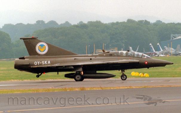 Side view of a military single engined jet fighter taxiing from left to right along a black runway.
The plane is in an olive green camouflage, with the white registration "OY-SKA" on the side of the rear fuselage, and a white, blue, and yellow crest on the tail.
The plane has a tandem 2 seat cockpit, one pilot sitting behind and above the other.
A black taxiway fills the foreground, with grass in the background leading up to trees in the distance, under grey sky.