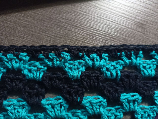 Detalle del borde de Emochal, apoyado sobre una mesa gris oscuro.
De abajo hacia arriba, se van alternando capas de verde azulado y negro, con grupos de 3 puntos altos y cadeneta entre grupos, para que el siguiente nivel construya sus grupos de puntos en ese hueco. Esto es el estilo granny.
La última capa, sin embargo, es una sucesión de puntos medios negros, sin hacer grupos ni dejar huecos.