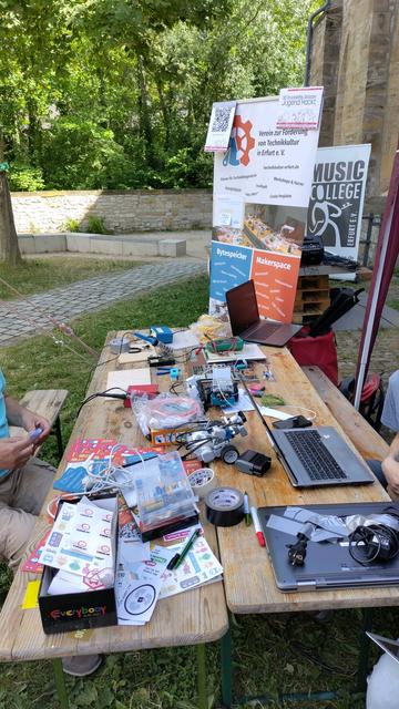 Auf zwei Biergarnituren befinden sich Laptops, Aufkleber, Flyer und Lötstationen. Hinter den Tischen steht ein Rollup-Banner des Bytespeichers und Makerspaces.