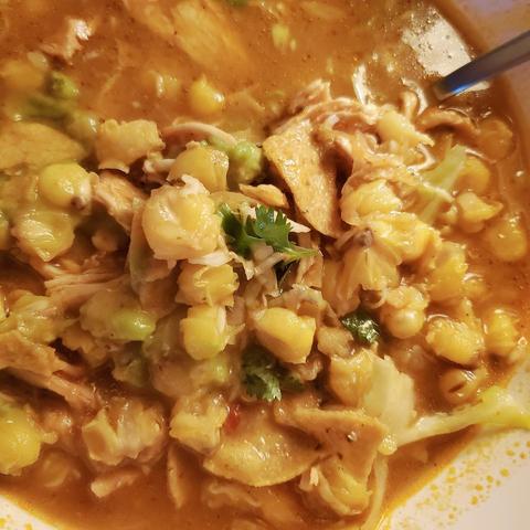 A close-up shot of a bowl of warm and comforting chicken pozole with tortilla chips soup. The soup contains hominy, shredded chicken, crisp tortilla strips, avocado, and shredded cabbage in a rich, slightly spicy broth. The soup is garnished with fresh cilantro. A spoon is visible in the upper right corner. The bowl is white with a hint of yellow.