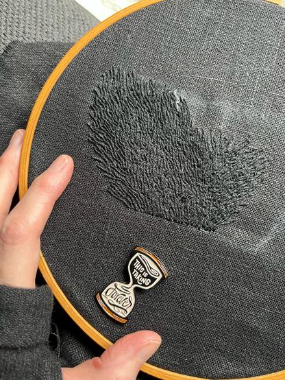 Lia’s fingers show a 4 by 5 inch wonky blob of black on black whipped backstitch that looks a bit like flames, but if you’ve been following this piece you’ll know it’s pubic hair. It’s definitely pubic hair. An hourglass-shaped needle minder that reads “this is taking forever” is attached to the cloth.