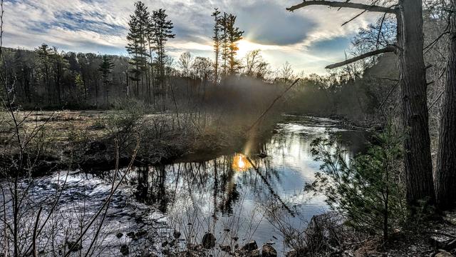 A very soft blue evening sky with lots of haze and clouds. Standing on a rocky slope just above the rocky bank of a small river. It is early spring, the grassy meadow across the river is dormant and golden brown. The hardwood trees and most shrubs still lack leaves. At the far edge of the meadow about a dozen towering pines stand at the near edge of a mixed forest of pines and leafless hardwoods. A low wooded ridge rises along the horizon to our left. To our right are the steep, wooded lower slopes of a mountain. The setting sun is just emerging below the clouds as a bright gold and orange orb mirrored on the river below.