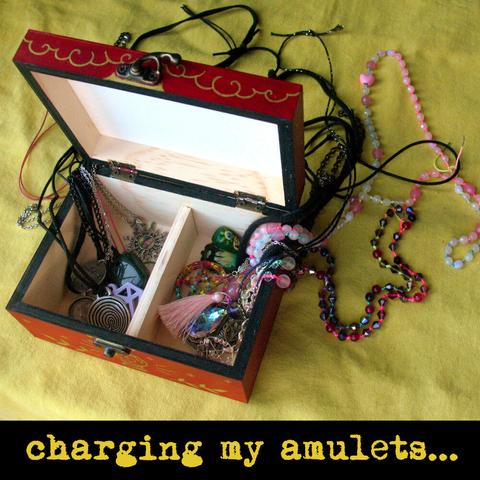 Red painted wooden box with the lid pen and two compartments inside. The chains of necklaces with amulets are coming out of the box and laying on a yellow textile surface. Text says: "charging my amulets...".