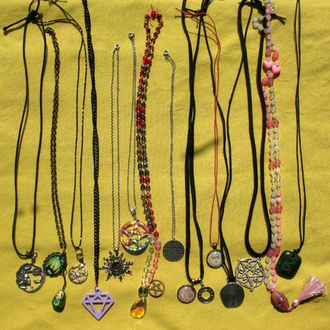 14 amulets and necklaces laying next to each other on a yellow textile surface. Among them are multiple pentagrams, lunar & solar amulets, references to witches and to Medusa, and two beaded necklaces.