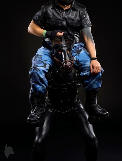 Against a stark black background, a person in full pony gear carries a rider on their shoulders. The pony, whose powerful thighs are clad in skintight, shiny black pants, wears a black leather corset-style harness. Their head is enclosed in a black leather horse hood, complete with a muzzle and bit, with only their eyes visible through the openings.

The rider, seated securely on the pony's shoulders, wears blue camouflage pants and black combat boots. Their upper body is dressed in a short-sleeved black leather shirt and a collar with a silver O-ring. One of the rider's hands, emerging from a sleeve that reveals a black forearm tattoo, grips the top of the pony's hood. Their other hand rests gently on the pony's knee. The dramatic studio lighting casts highlights on the supple leather and the defined muscles, creating a powerful and sensual scene.
