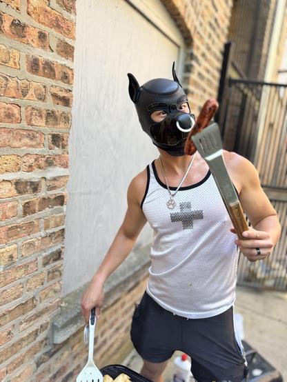 The image focuses on the pup in the camo/black hood, who playfully sticks his tongue out while holding a sausage aloft on his tongs. A third photo shows a shirtless pup in a black hood squatting down, intently flipping burger patties on the gr