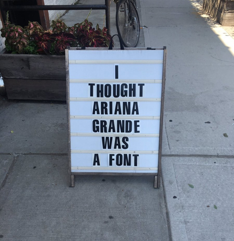White sandwich board sign on a sidewalk with black text reading:
"I THOUGHT ARIANA GRANDE WAS A FONT".
