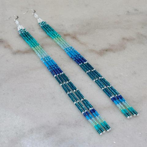 A pair of long beaded dangle earrings in shades of blue, turquoise, and white, on a marble background.