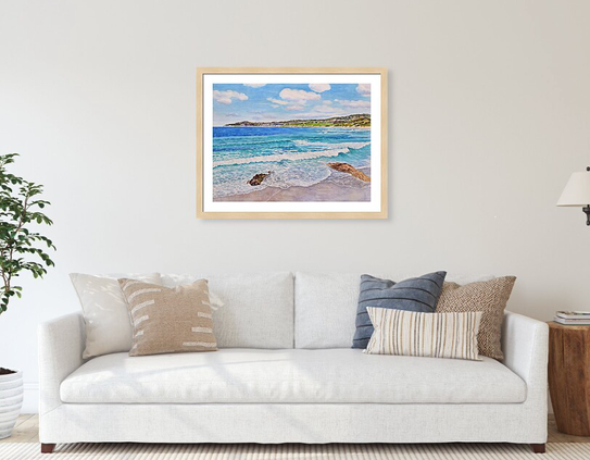 A watercolor painting hangs on a wall above a couch with pillows. The painting is a beach scene with blue and turquoise colors.