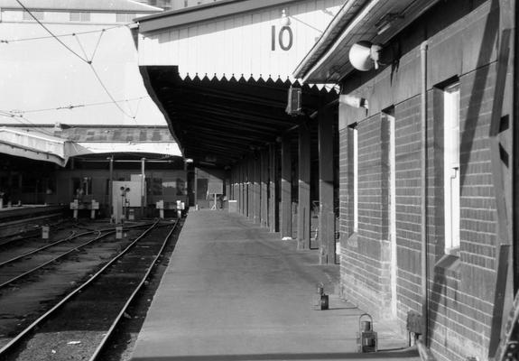 A black and white photo taken looking along a passenger platform at a city terminal station.  Tracks are at the left with buffers at the end in front of the transverse concourse behind.  At right, a wooden awning covers the platform on which we are standing.  The end of the awning is decorated by downward pointing pickets or daggers, and a large number 10 is displayed on the fascia to denote the platform number.  At bottom right, two portable lamps are sitting on the platform.  Lamps like this, fuelled by oil, paraffin or kerosine, were used from the dawn of railways until the early 1970s.  Were oil lamps still really in use in 1985 - or were these perhaps relics?  Could these have been more modern battery electric versions?