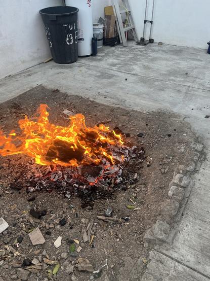 A small fire burning on the ground, surrounded by ashes and debris. In the background, there is a trash can, a ladder, and various containers. The area appears to be an outdoor space with concrete flooring.