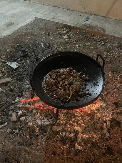An oversized cast iron pan sits over an open fire, containing cooking food items. The surrounding area is dirt and ash, with scattered debris. The scene is illuminated by the glow of the embers beneath the pan.