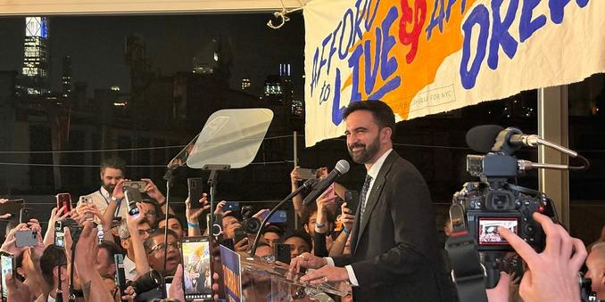 Zohran Mamdani stands at a podium on a stage before many people. The people hold up phones and cameras and microphones. A banner with a portion of some campaign slogan is raised behind him but the slogan is not readable