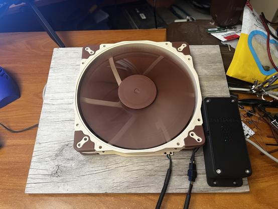 A brown 7” fan mounted on a piece of plywood with a small black enclosure next to it. The fan is running and green lights can be faintly seen through the vent holes of the black box.