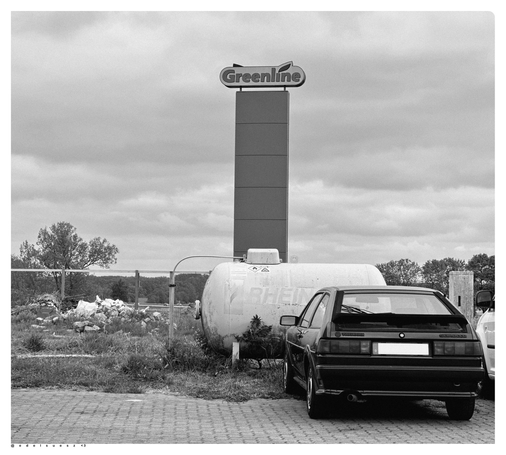 Schwarzweißfotografie: an einer Tankstelle fotografiert, links Bauschutt auf einer Wiese, daneben eine große Werbetafel der Tankstelle, Aufschrift „greenline“, davor ein weißer Gastank und ein alter abgestellter Volkswagen SCIROCCO von hinten