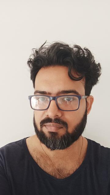 A man with rectangular specs looking in the camera. 

He has short somewhat wavy hair. 

There's a beard and a moustache.

Wearing a dark blue tee, and a chain in the neck.
