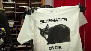 A YouTube thumbnail featuring a man in a cluttered repair shop obscuring his face and torso by holding up a T-shirt with a picture of a very angry black cat and the text “SCHEMATICS OR DIE”. He’s wearing latex gloves.
