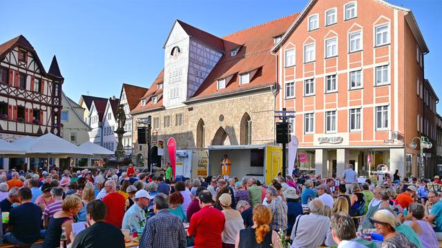 Bei wolkenlosem Himmel sitzen hunderte Menschen an Biertischgarnituren auf einem Platz im Schatten. Auf den Tischen stehen Blumen, Getränkeflaschen und Plastikschüsseln. Auf einem Anhänger steht eine Frau und spricht in ein Mikrofon.