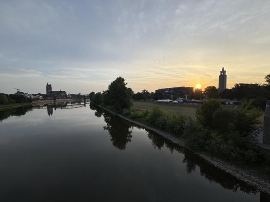 Am Horizont eine Kirche (Magdeburger Dom), im Vordergrund, ein nahezu spiegelglatter Fluss (die Elbe) darüber die aufgehende Sonne gleich rechts hinter einem großen Gebaude (Stadthalle und Baustelle), der Fluss ist gesäumt von grünen Bäumen, Büschen und Wiesen, die Stadtsilhouette von Magdeburg über die Elbe Richtung Dom geblickt am frühen Morgen