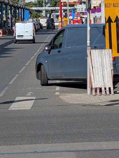 Foto einer zweispurigen Straße, die rechts einen sehr schmalen aufgemalten Radstreifen hat. Ein dunkler Lieferwagen parkt halb auf einem Parkplatz und halb am Radstreifen und versperrt ihn damit.