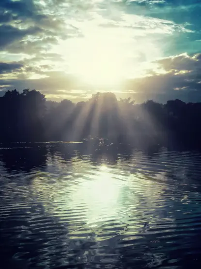 Das Bild zeigt einen Sonnenuntergang über einem ruhigen Gewässer.  Die Sonne scheint hinter einer Baumlinie hervor und erzeugt strahlende Licht-Reflexe auf dem Wasser. Unterhalb der Baumlinie ist schemenhaft ein Floß oder Boot zu sehenn. Der Himmel ist mit Wolken bedeckt, die in verschiedenen Blautönen und Gelb erscheinen. Das Wasser ist ruhig, aber mit leichten Wellenmustern, die das Licht reflektieren. Die Gesamtstimmung ist friedlich und ruhig.