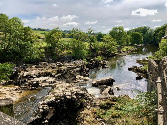 This picture captures a serene riverside scene, characterized by clear, gently flowing water surrounded by lush greenery. The river meanders through rocky terrain, with large, rugged stones and boulders breaking the water's surface. On both sides of the river, dense trees with vibrant green foliage stretch out, indicating a healthy, natural environment. The sky above is partly cloudy, allowing beams of sunlight to illuminate parts of the landscape, creating a lively contrast between light and shadow. A stone wall or structure is visible on the right side, adding a touch of human-made elements to this otherwise natural setting.