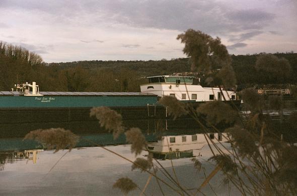 Colored photography of a barge (from the middle to the rear cabin) on the river, with reflection and plants in the foreground. The sky is grey abd cloudy.