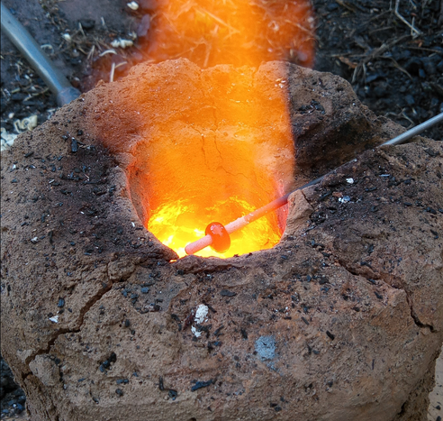 Die heiße Flamme aus der Ofendüse erhitzt einen hinein gehaltenen Dorn mit einer aufgewickelten Glasperle, die glüht und nun rund gedreht wird.