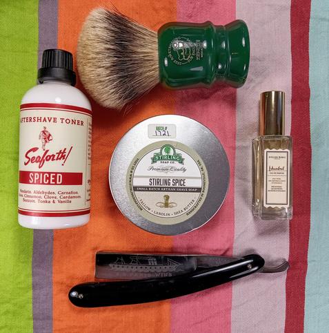 (Le Chat Mistral description)

The image showcases a collection of grooming items arranged on a striped background. At the top left, there's a bottle of Seaforth Spiced Aftershave Toner. Next to it, towards the centre, is a tin of Stirling Soap Co. Stirling Spice shaving soap. Above the soap, there's a shaving brush with a green handle. To the right of the brush, there's a small bottle of Istanbul Eau de Parfum. At the bottom of the image, there's a black-handled straight razor with "S.S. Paris Wins" engraved on the blade. The items are neatly laid out, suggesting a traditional shaving setup.