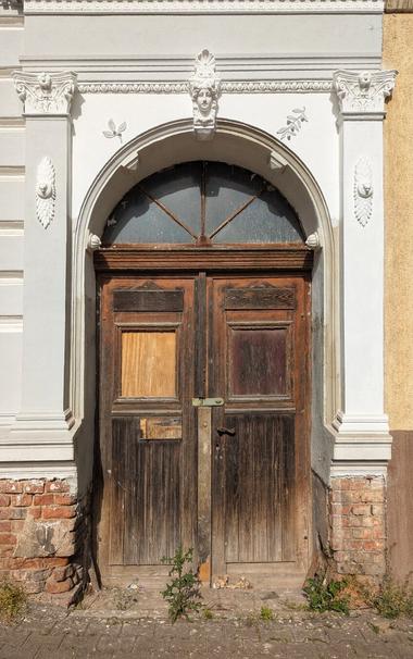A weathered, dark wooden double door with an arched transom window above it is set into a grand, ornate white architectural surround. The door shows signs of age with faded paint and exposed wood grain. One of the rectangular panels on the left door is covered with a lighter, possibly plywood, patch. A simple metal latch is visible where the two doors meet.
The white door frame features decorative elements including a central sculpted face at the top of the arch, flanked by scrollwork and floral motifs. Two substantial columns with Corinthian-style capitals frame the door, supporting a detailed entablature. Below the white frame, the lower part of the wall is exposed red brickwork, showing some deterioration. The ground in front of the door is a paved surface with some weeds growing through cracks, and to the right, a plain, light brown wall is visible.