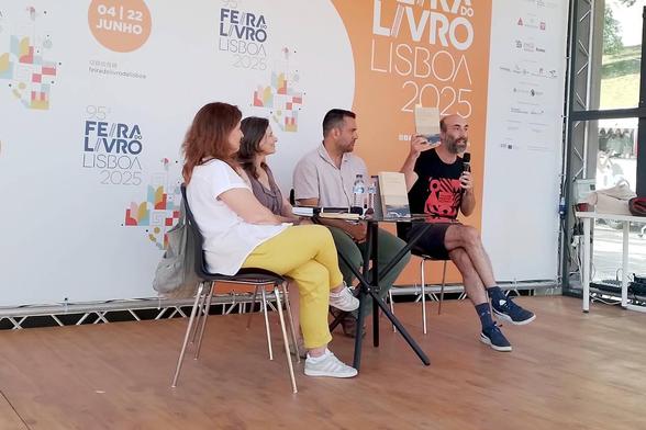Elisa Lopes da Silva, Cláudia Castelo, Ricardo Noronha e Victor Pereira sentados no palco da Praça Laranja da Feira do Livro de Lisboa. Victor Pereira está a falar e tem o livro "Colonizar a Metrópole", de Elisa Lopes da Silva, erguido na mão. A fotografia foi tirada por Catarina Laranjeiro.