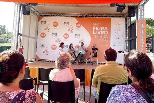 Elisa Lopes da Silva, Cláudia Castelo, Ricardo Noronha e Victor Pereira sentados no palco da Praça Laranja da Feira do Livro de Lisboa. Ricardo Noronha está a falar. A foto foi tirada por Rita Luís a partir do público, que está sentado de costas para a câmara.
