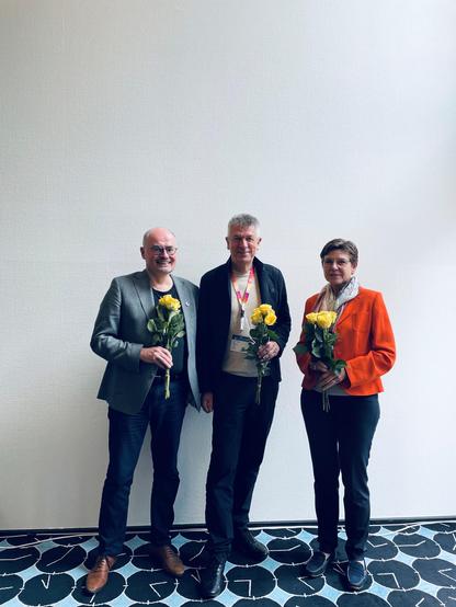 Drei Personen stehen vor einer weißen Wand und halten gelbe Rosen in der Hand. Sie lächeln in die Kamera.