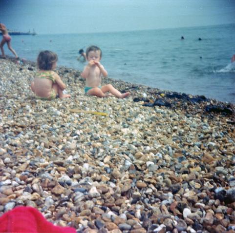 A scan of a square format, colour photograph taken in the early 1970s. We're on a pebble beach with part of a red blanket visible in the foreground low down, and we're looking across a short stretch of pebbles towards the relatively calm sea as it laps against the gently sloping mass of small, smooth stones. The angle of the shot is slightly off so the horizon is slightly tilted down to the left and in the distance we can see the end of a pier sticking out. There are heads of people swimming in the water and a couple of children are coming out of the sea in the background, one wearing a scuba mask. The main focus is on two children aged no more than 2 or 3 sat on the pebbles a couple of metres in front. A boy is turned, facing a little towards our view and is peering intently at a pebble or shell in his hands. A girl squats next to him with her back to us. The colours are faded but it feels like a warm day and there's clearly very little wind given the sea conditions.