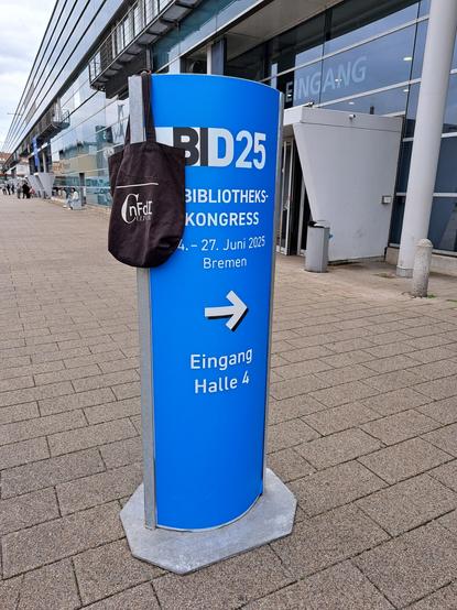 An einem vertikalen Hinweisschild vor der Veranstaltungshalle des Bibliothekskongresses (BID25, BiblioCon25), der vom 14. bis 27. Juni in Bremen stattfindet, hängt ein NFDI4Culture-Beutel.