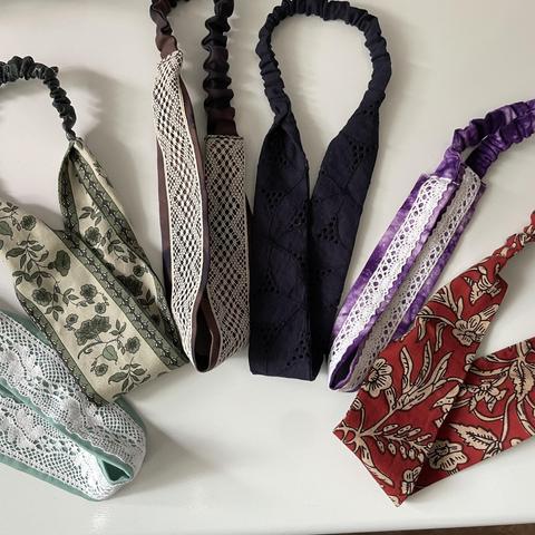 Six hairbands on a white surface. From left to right: mint green with a white lace, green printed pattern, tie dye brown with a beige lace, navy blue, tie dye purple with a white lace, red printed pattern. The hairbands all have elastic ribbon on one end.