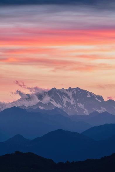 A majestic mountain range is depicted under a colorful sunset sky, featuring shades of pink and purple. Clouds hover around the peaks, with darker mountains in the foreground creating a layered effect.