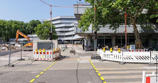 Baustelle vor dem Philosophikum mit Absperrungen und einem orangefarbenen Bagger, der im Hintergrund arbeitet.