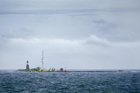 A small island in the sea with a lighthouse and some communications building with an antenna. Lower part of the photo is just a wavy sea and the upper three fourths is clouds in the background