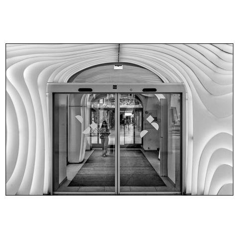 A black and white picture of a modern shopping mall entrance featuring flowing, curved white walls with horizontal ribbed patterns that create a tunnel-like effect around glass doors. A silhouetted figure walks through the bright interior space beyond the entrance.