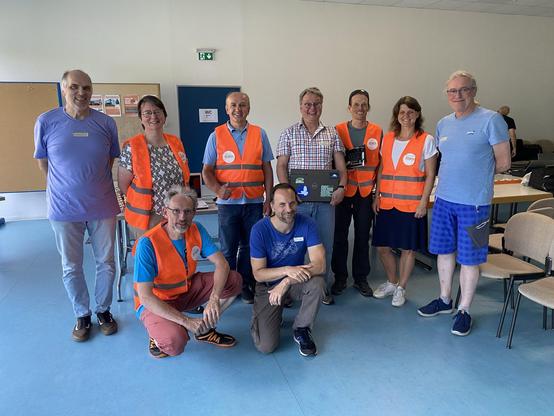Gruppenfoto des Organisationsteams im Übungsraum des Bürgerhauses, bestehend aus sieben Männern und zwei Frauen. Darunter sind fünf Mitglieder des Bürgerausschusses, die an orangefarbenen Warnwesten mit einem Aufkleber zu erkennen sind. Alle lächeln in die Kamera.