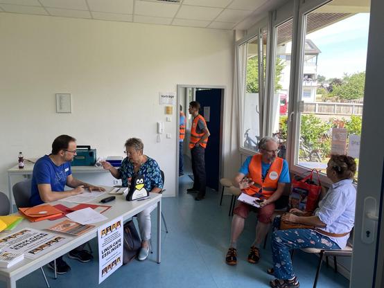 Blick in den Empfang: links sprechen ein Mann hinter einem Schreibtisch und eine Frau daneben sitzend miteinander. Der Tisch ist bedeckt mit Unterlagen und Formularen. Daneben sitzen sich ein Mann und eine Frau auf Stühlen gegenüber und reden miteinander. Im Hintergrund sind durch eine offene Tür zwei weitere Personen zu sehen, die stehend einen Vortrag im Vortragsraum verfolgen.