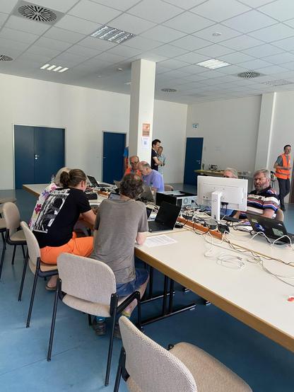 Das Bild zeigt eine Gruppe von Menschen, die in einem hellen, modernen Raum an einem langen Tisch sitzen und an Computern arbeiten. Der Raum hat weiße Wände und ein blaugrünes Bodenlaminat. An der Decke sind quadratische, graue Kacheln angebracht, die mit fluoreszierenden Leuchten ausgestattet sind. Auf dem Tisch sind mehrere Laptops und Monitore zu sehen, zusammen mit Kabeln und anderen elektronischen Geräten. Einige Personen sind konzentriert an ihren Geräten arbeitend, während andere miteinander sprechen. Im Hintergrund sind mehrere Türen zu sehen, die in einem dunklen Blau lackiert sind. Einige Personen tragen leichte, sommerliche Kleidung, wie T-Shirts und Shorts, was auf eine informelle Atmosphäre hinweist. Ein Mann in einem orangefarbenen Warnweste steht am Ende des Raumes und beobachtet die Gruppe. Die Szene wirkt produktiv und kooperativ, mit einer Mischung aus Konzentration und Gespräch.