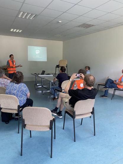 Das Bild zeigt eine Gruppe von Menschen in einem einfach eingerichteten Raum, der wie ein Konferenzraum aussieht. Im Vordergrund sitzen mehrere Personen auf beigen Stühlen, die auf eine Person in einem orangefarbenen Warnweste zuzuschauen scheinen, die am Kopfende des Raumes steht und eine Präsentation hält. Auf einem Projektionsbildschirm hinter dem Redner ist der Text "Vielen Dank" und ein Logo mit einem "M" zu sehen. Einige Zuhörer nehmen Notizen oder fotografieren die Präsentation mit ihren Smartphones. Die Wände sind weiß, der Boden ist blau, und die Decke ist mit grauen Kacheln verkleidet. Die Atmosphäre wirkt professionell und informell zugleich.