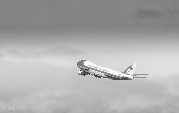 This is a black and white aviation photo is Air Force One in flight. In the lower right corner is an oblique view, with the left side and rear in sight, of the Boeing 747 used by the president of the USA. The aircraft is flying from right to left and away from the camera. On the top of the white fuselage, in large bold letters the words: United States Of America with a Large stars and stripes flag on the vertical fin. While the aircraft is in the clear it's surrounded by distant clouds.
If you were curious the passenger was the 45th president